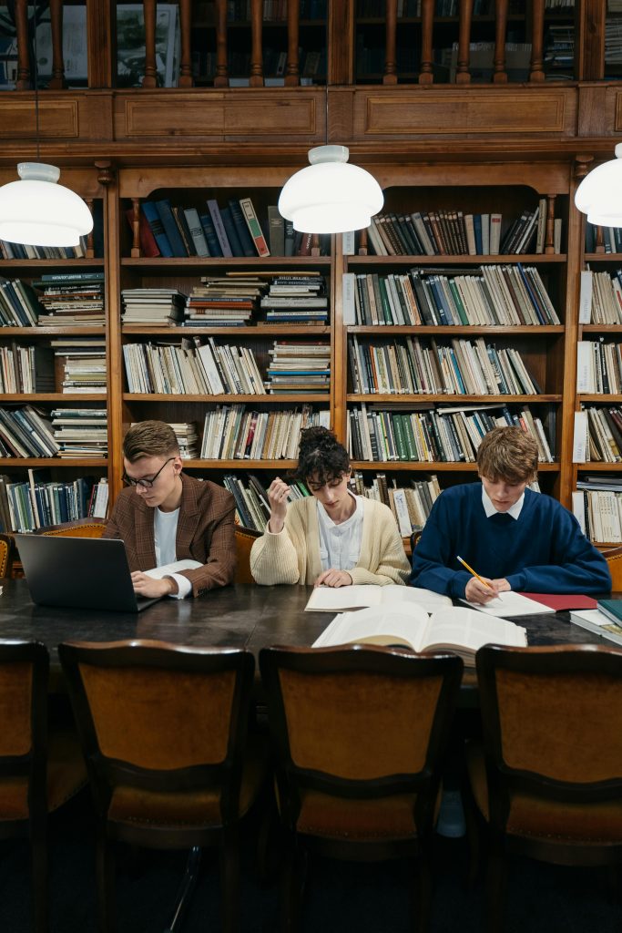 College students engaged in study session at a library table surrounded by shelves of books.